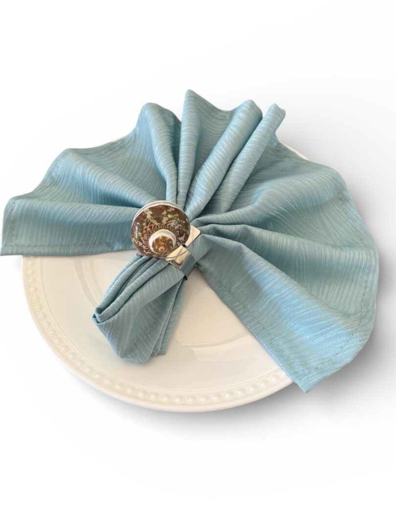 Light blue cloth napkin with a brown flower napkin ring on a white plate.